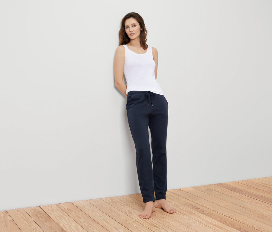 Eine Frau lehnt an einer Wand und trägt ein weißes Tanktop und eine marineblaue Trainingshose.