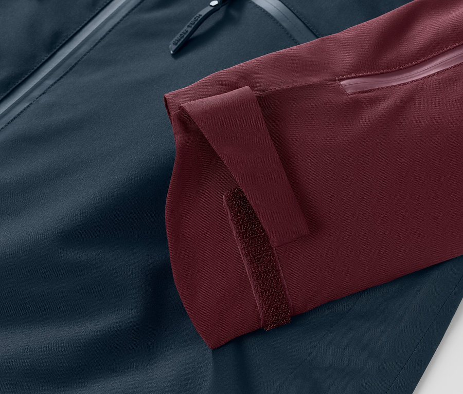 Detailaufnahme einer Ski-und-Snowboardjacke: bordeauxroter Ärmel mit verstellbarer Klettverschlussmanschette und marineblauer Rest der Jacke.