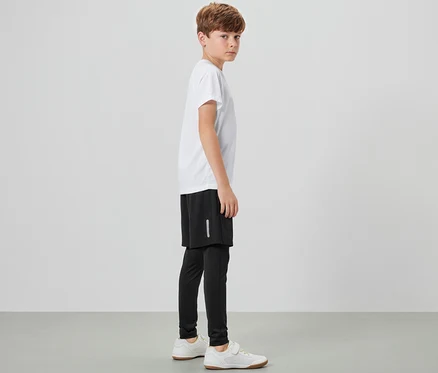 Ein Junge steht seitlich und trägt eine Kinder-Sporttight mit Shorts, ein weißes T-Shirt und Turnschuhe.