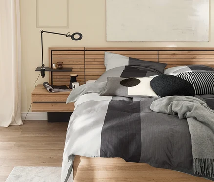 Modernes Schlafzimmer mit Holzbett und grauer gestreifter SCHÖNER WOHNEN-Kollektion ART Bettwäsche, Zierkissen und einer grauen Decke. Eine schwarze Lampe ist an einem Holznachttisch neben dem Bett befestigt.