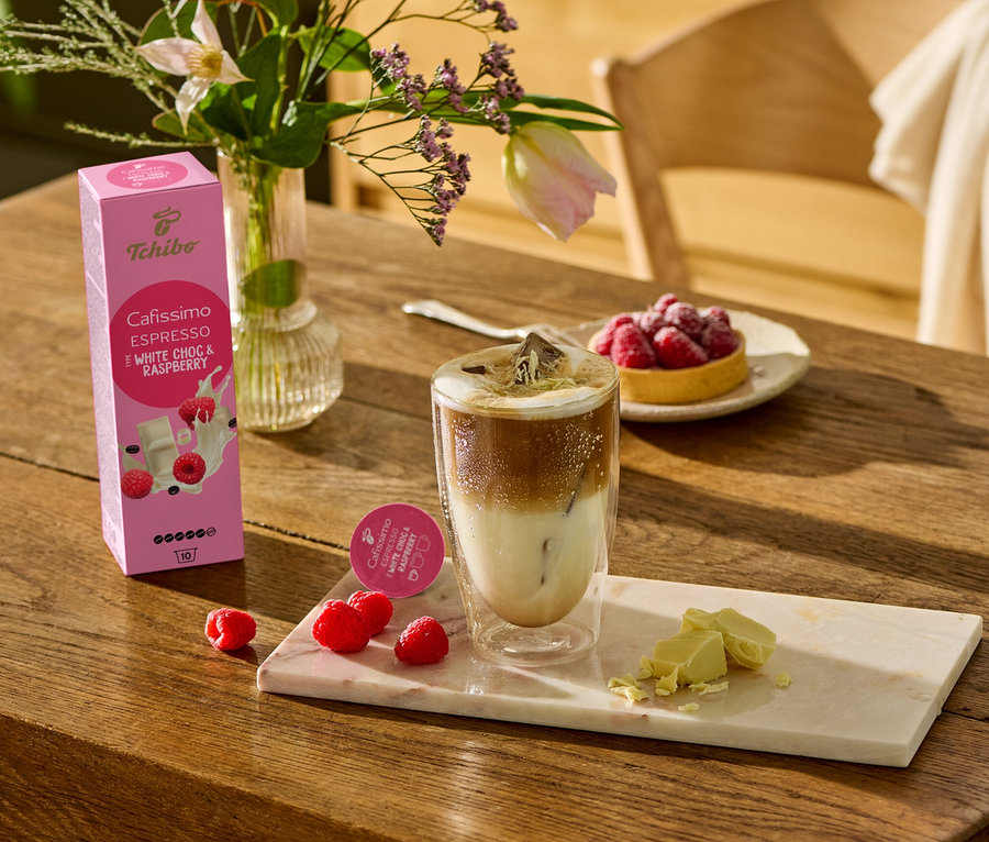 Auf einem Holztisch stehen ein Glas Kaffee, weiße Schokoladenstücke und Himbeeren auf einem Marmorbrett. Im Hintergrund ist eine Packung Cafissimo Flavoured Espresso - White Choc & Raspberry - 10 Kapseln zu sehen.