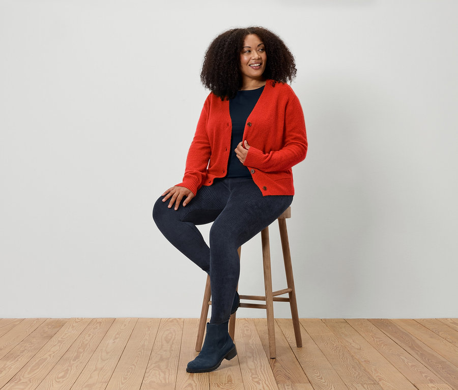 Eine Frau sitzt auf einem Hocker und trägt eine rote Strickjacke, blaue Jeans und blaue Stiefel.