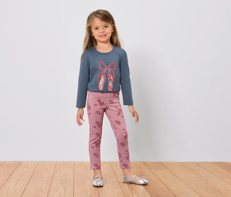 Mädchen im blauen Langarmshirt mit Ballettschuhen und rosa Leggings steht auf einem Holzboden.
