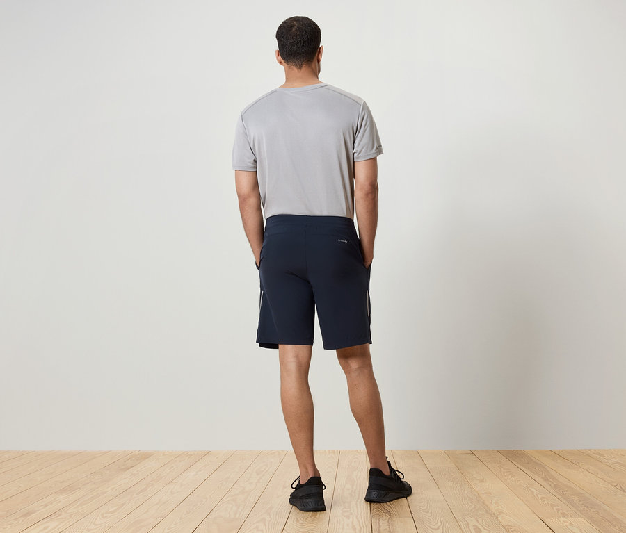 Ein Mann mit dem Rücken zur Kamera trägt ein graues T-Shirt, dunkelblaue Shorts und schwarze Turnschuhe.