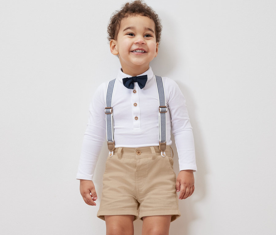 Lächelnder kleiner Junge in weißem Langarmhemd mit Fliege, Hosenträgern und Kinder-Shorts.