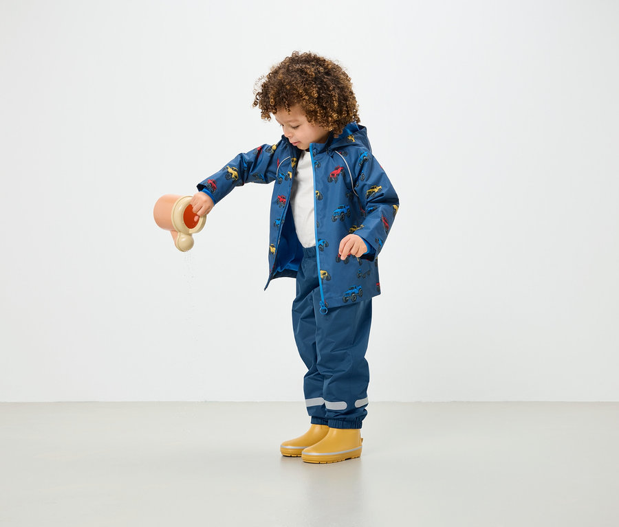 Ein Kind mit lockigem Haar gießt Wasser aus einem Spielzeug, während es einen blauen, bedruckten Regenmantel und blaue Hosen trägt.