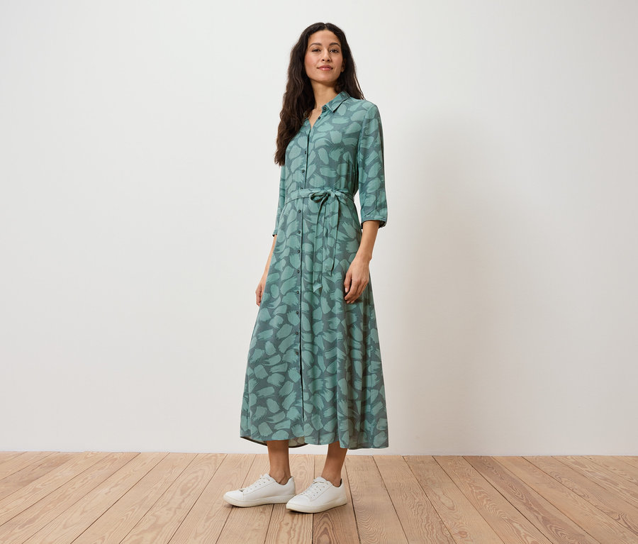 Eine Frau steht und trägt ein grünes Hemdkleid mit Aufdruck und weiße Turnschuhe.