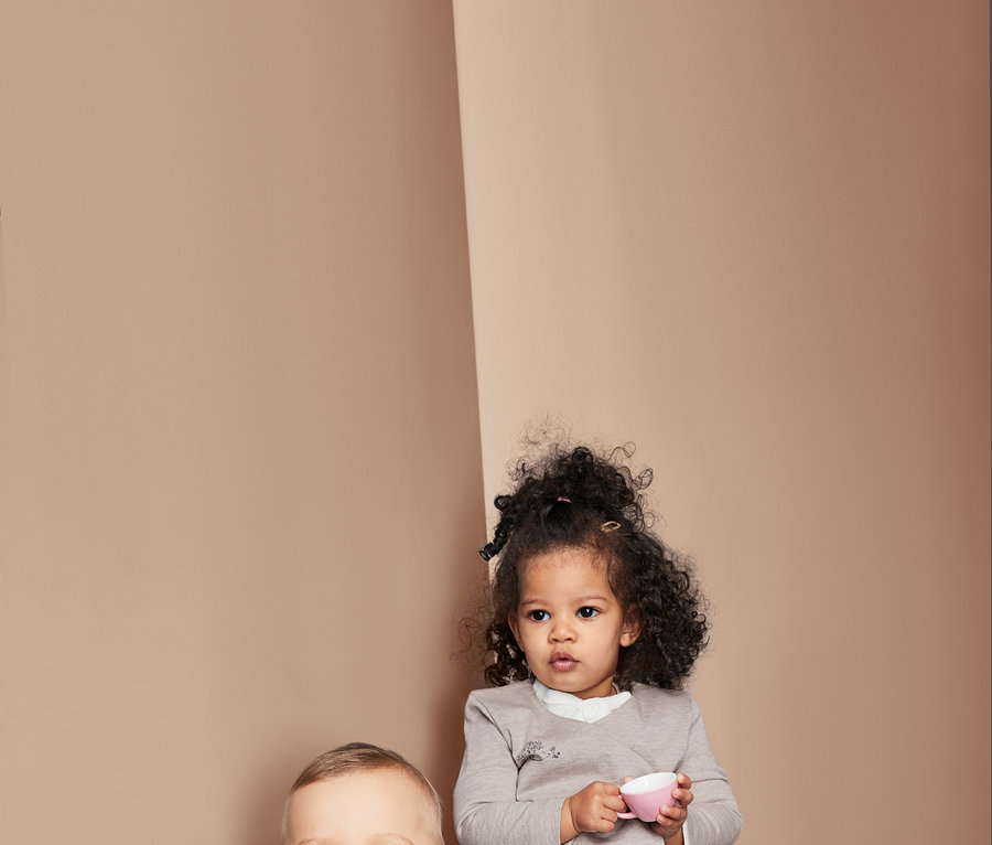 Zwei Kinder sitzen da. Ein Junge sitzt auf einem Kissen und lächelt, er trägt ein gestreiftes Shirt. Ein Mädchen hält eine rosa Tasse und trägt ein Oberteil. Beide tragen Jeans.