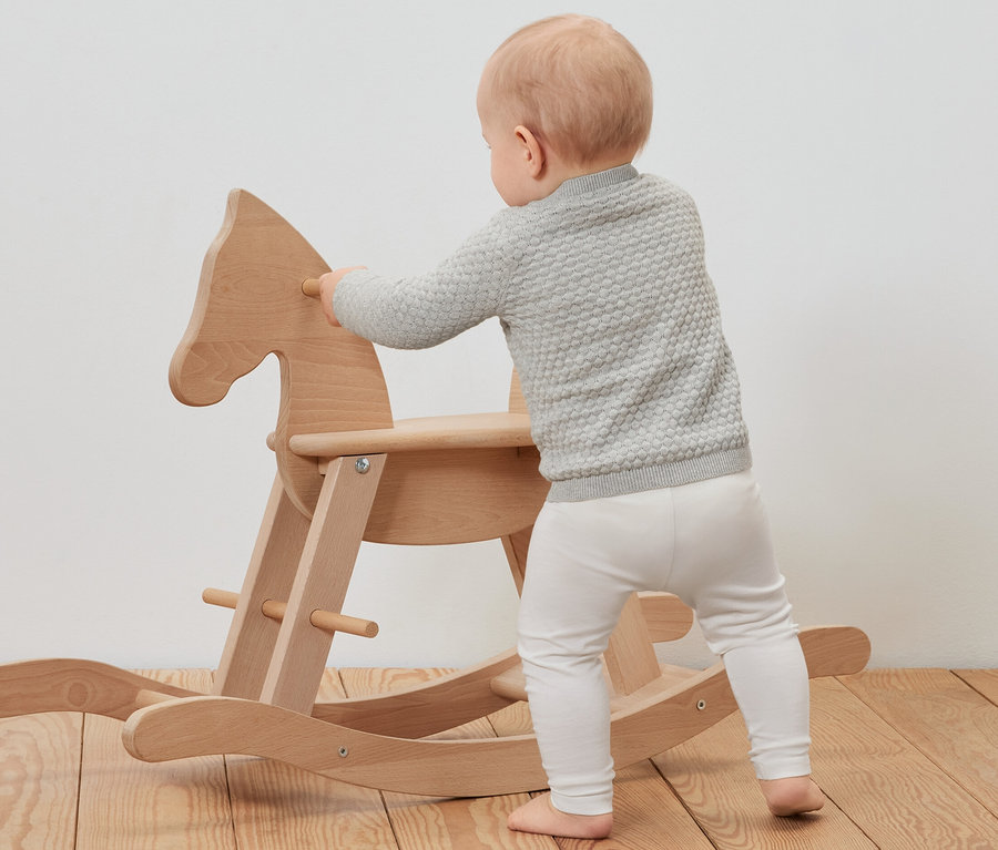 Kleinkind in Baby-Strickjacke lehnt sich an ein hölzernes Schaukelpferd.