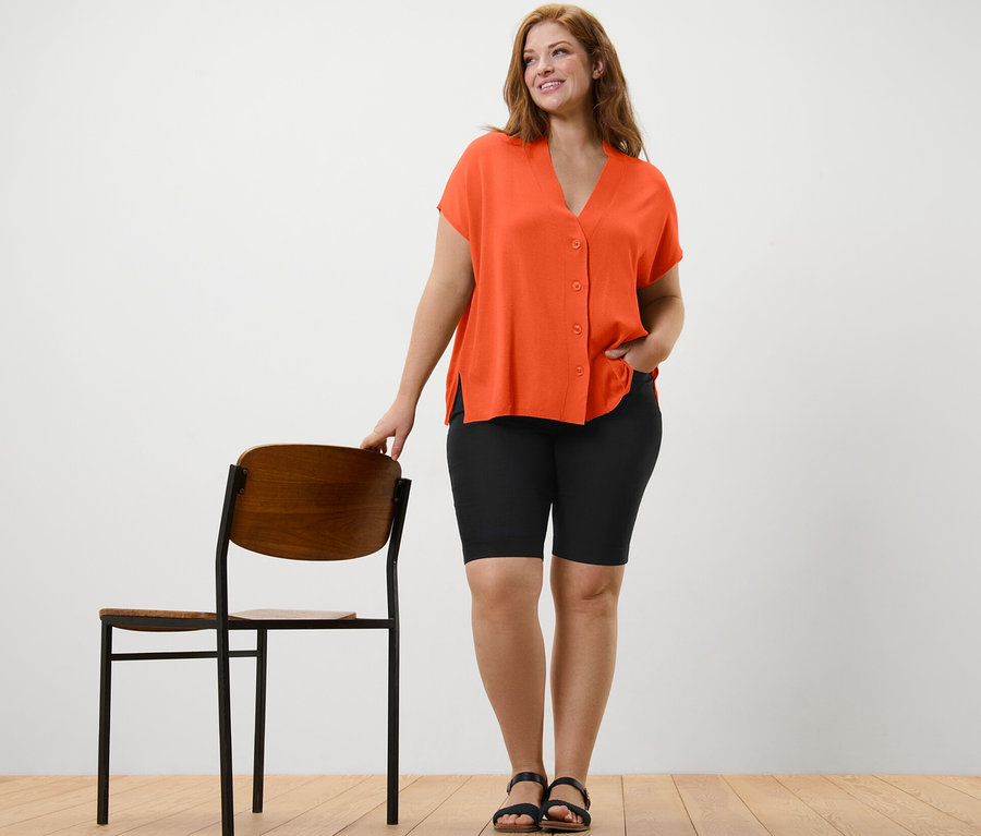 Frau steht neben einem Stuhl und trägt einen orangefarbenen Kurzarm-Cardigan und eine schwarze Bengalin-Bermudashorts.