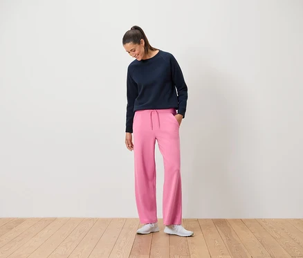 Eine Frau in einem marineblauen Sweatshirt und einer pinkfarbenen Hose steht auf einem Holzboden.