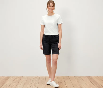 Frau steht auf Holzboden und trägt Bermuda Jeans ‒ Fit »Lea«, black denim und ein weißes T-Shirt.