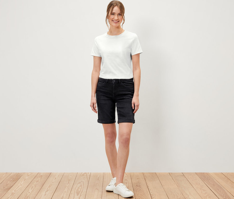 Frau steht auf Holzboden und trägt Bermuda Jeans ‒ Fit »Lea«, black denim und ein weißes T-Shirt.