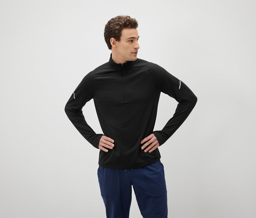 Mann mit braunen Haaren posiert mit den Händen in den Hüften, gekleidet in einem schwarzen Langarmshirt und blauen Hosen.