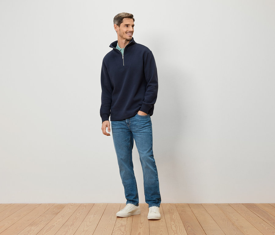 Ein Mann steht vor einer weißen Wand. Er trägt einen marineblauen Pullover mit Reißverschluss, blaue Jeans und weiße Schuhe.