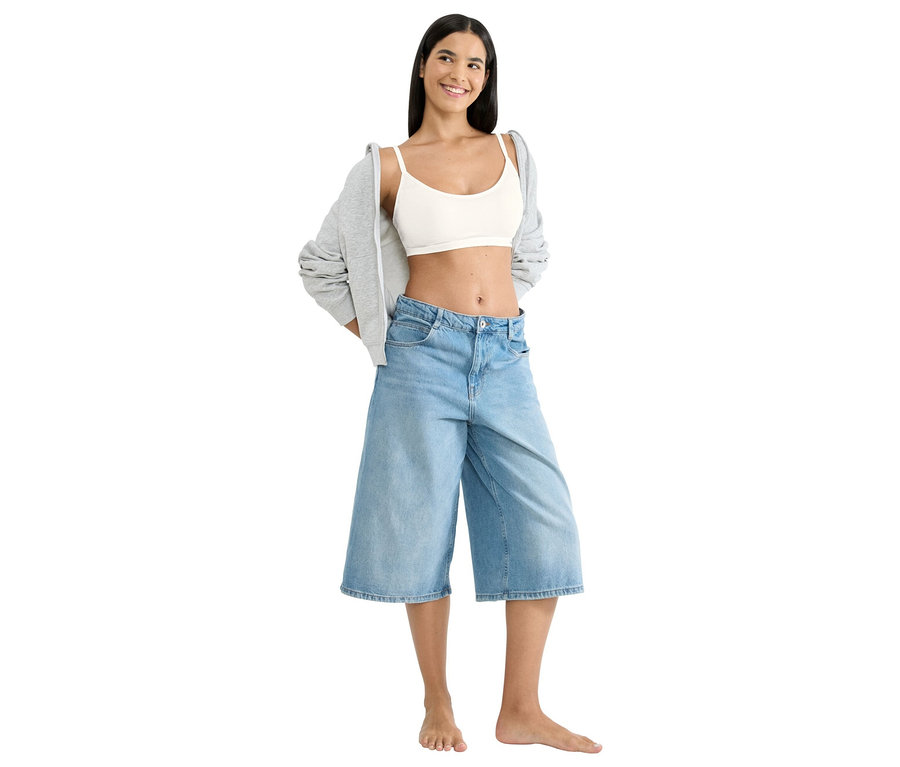Eine lächelnde Frau mit dunklem Haar steht barfuß und trägt einen weißen sloggi Free Evolve P BH, hellblaue weite Jeans und einen grauen Hoodie über den Schultern.