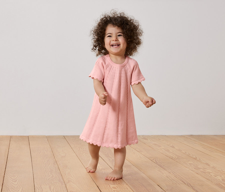 Lachendes Kleinkind mit lockigem Haar, das ein rosa Kleid mit gewelltem Saum auf einem Holzboden trägt.