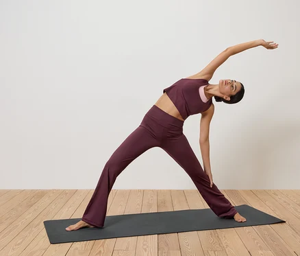 Frau in Burgunder Sportbekleidung dehnt sich auf einer Yogamatte auf einem Holzboden vor einer weißen Wand.