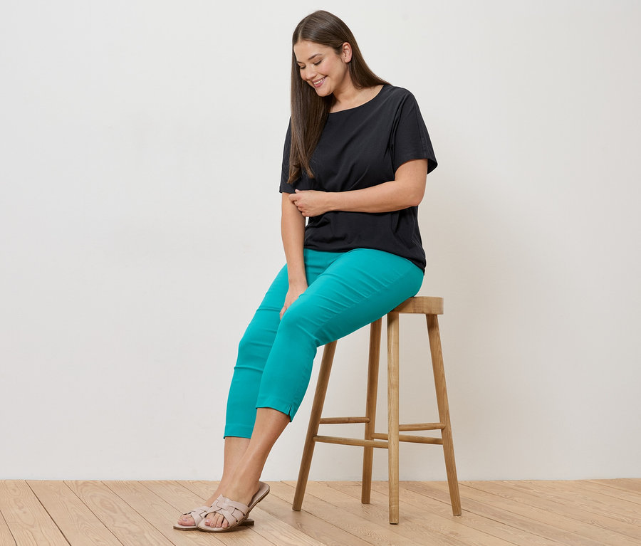 Frau sitzt auf Hocker, trägt schwarzes Blusenshirt und azurblaue 7/8-Stretchhose.