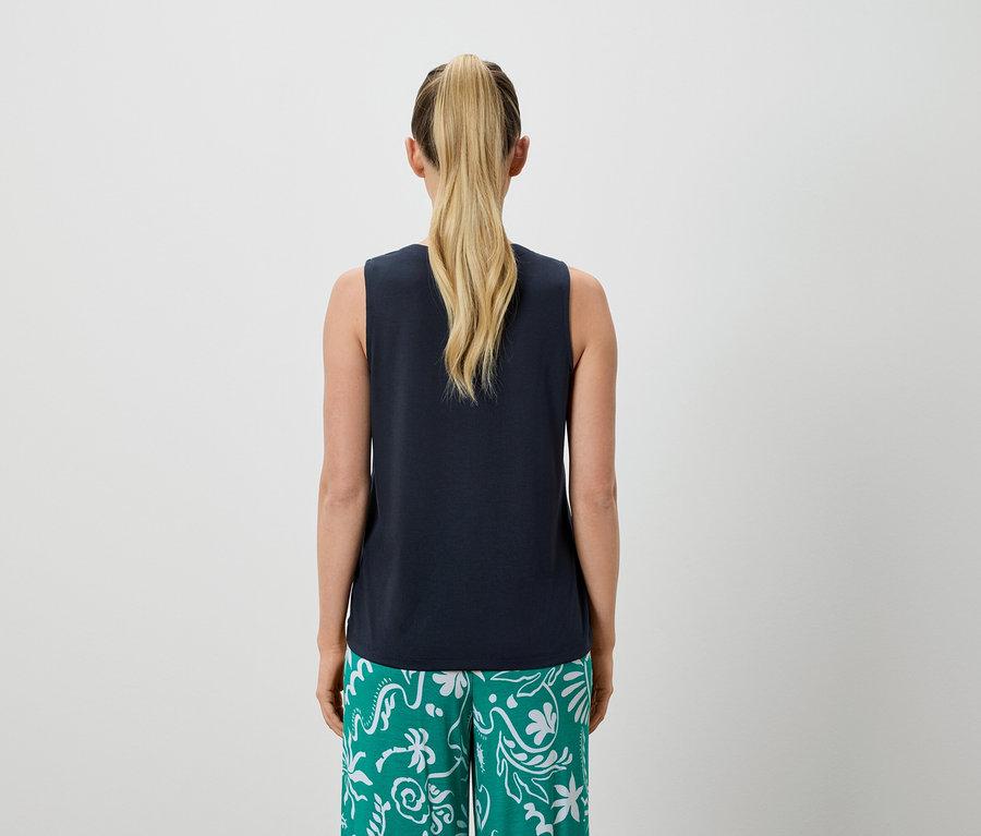 Rückansicht einer Frau mit blondem Pferdeschwanz, die ein marineblaues Tanktop und grüne, gemusterte Hosen trägt.