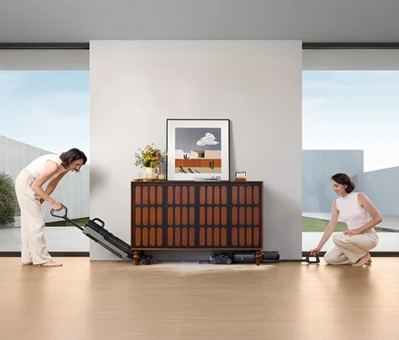 Zwei Frauen benutzen den Tineco Nass-Trockensauger »S7 Stretch Steam Plus« auf einem Holzboden vor einem Sideboard.