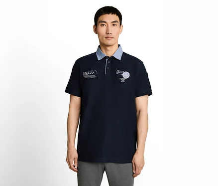 Ein Mann mit asiatischem Aussehen trägt ein dunkelblaues Poloshirt mit hellblauem Kragen, weißen Aufdrucken auf der Brust und grauer Hose.