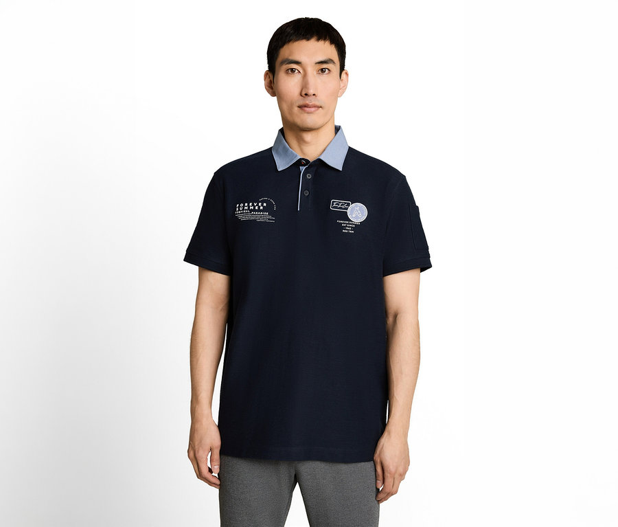 Ein Mann mit asiatischem Aussehen trägt ein dunkelblaues Poloshirt mit hellblauem Kragen, weißen Aufdrucken auf der Brust und grauer Hose.
