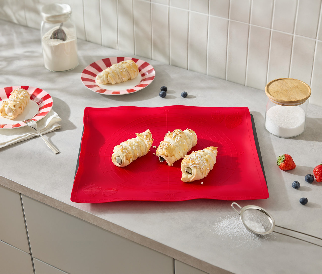 Drei mit Creme gefüllte Hörnchen liegen auf einer roten Unterlage, umgeben von Zutaten wie Erdbeeren und Blaubeeren.