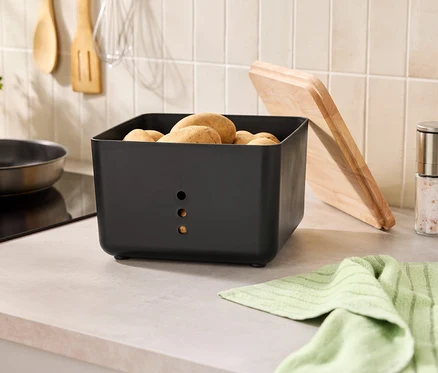 Auf einer Küchentheke steht ein schwarzer Kartoffelbehälter mit Kartoffeln, daneben ein Schneidebrett, Gewürzmühlen und ein Geschirrtuch.