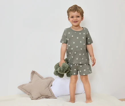 Ein kleiner Junge steht barfuß auf einem Bett mit einem olivgrünen Sternen-Pyjama und hält ein grünes Stofftier.