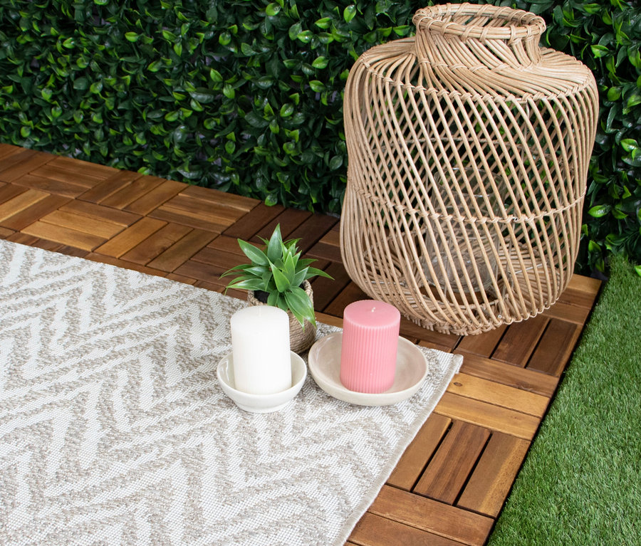 Outdoor-Teppich »Ecru Flow« mit hellem geometrischem Muster auf Holzfliesen, umgeben von Kunstrasen und einer grünen Hecke. Darauf stehen eine weiße und eine pinke Kerze in Schalen, eine kleine Topfpflanze und eine große geflochtene Rattan-Laterne.
