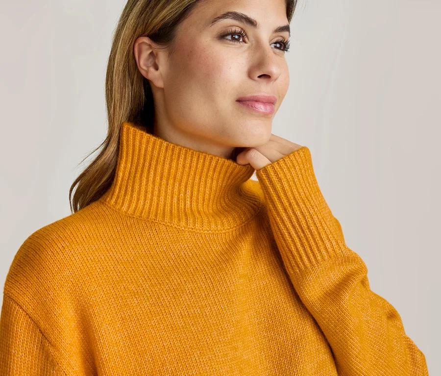 Nahaufnahme einer Frau in einem senfgelben Rollkragenpullover, die nach rechts blickt und sich sanft ans Kinn fasst.