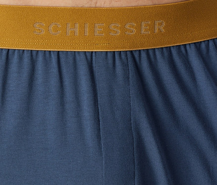 Nahaufnahme von blauem Stoff mit einem goldenen Gummibund, auf dem der Markenname „SCHIESSER“ in perforierten Buchstaben zu sehen ist.