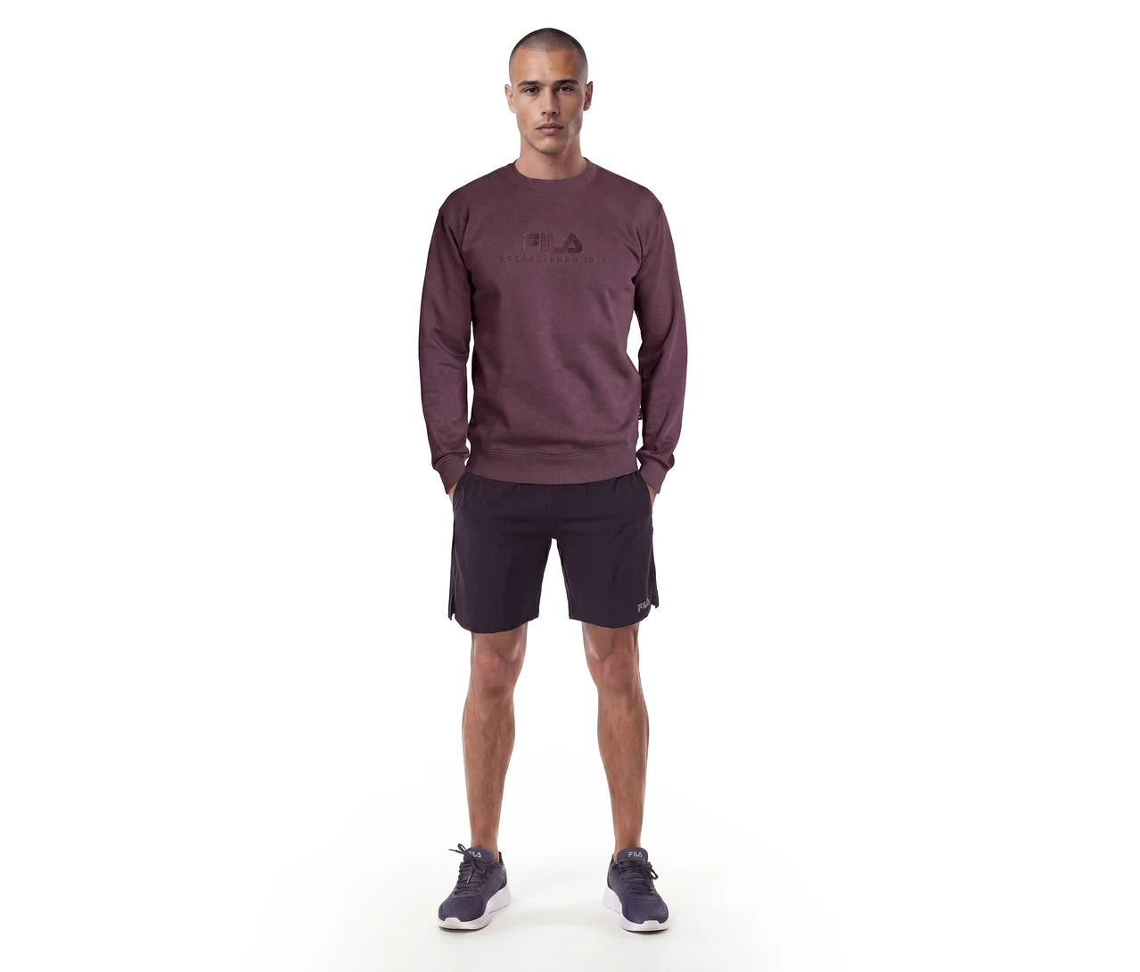 Mann steht in einem dunkelroten Sweatshirt mit Fila-Logo, schwarzen Shorts und dunklen Turnschuhen.