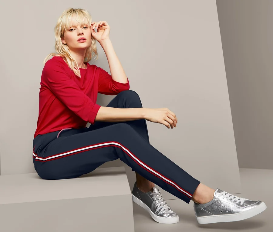 Blonde Frau sitzt und trägt ein rotes Shirt und eine Joggpants mit Galonstreifen.