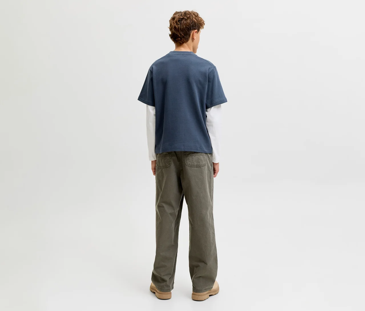 Eine Person steht mit dem Rücken zum Betrachter und trägt ein Jack&Jones double layered Shirt, bestehend aus einem blauen Kurzarmshirt über einem weißen Langarmshirt, dazu olivgrüne Hosen und hellbraune Schuhe.