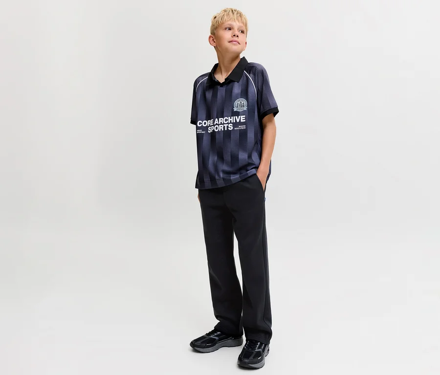 Blonder Junge in gestreiftem Poloshirt und schwarzen Jack&Jones Bradley Sweatpants. Er steht mit den Händen in den Taschen und blickt nach rechts.