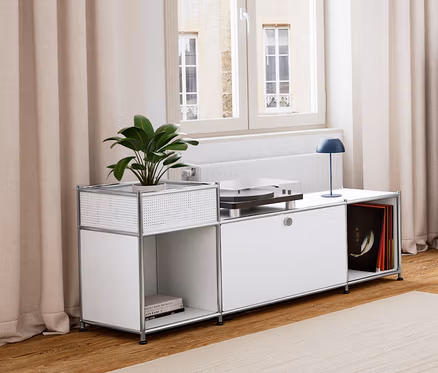 Ein Wohnzimmer mit Holzboden und hellem Teppich. Links steht ein weißes Lowboard Metall »CN3« mit Klappenfach, darauf eine dunkelblaue LED-Akku-Tischleuchte. Daneben eine dunkelblaue Kommode Metall »CN3« mit 3 Klappenfächern. In der Mitte ein Beistelltisch mit Tablett und Sitzfunktion und ein Sessel. Im Hintergrund ein hellgraues Sideboard Metall »CN3« mit 3 Klappenfächern.