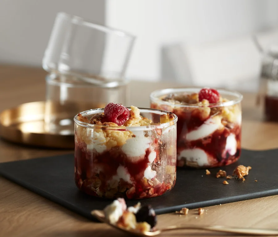 Auf einem Tisch stehen 4 Portions-und-Ofengläser, gefüllt mit einem Fruchtdessert. Zwei Gläser stehen auf einem schwarzen Teller, zwei weitere sind auf einem goldenen Tablett gestapelt.