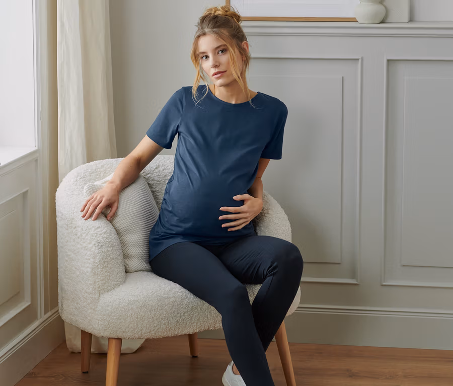Schwangere Frau sitzt im Sessel und trägt ein blaues Umstands-Shirt aus einem 2er-Set.