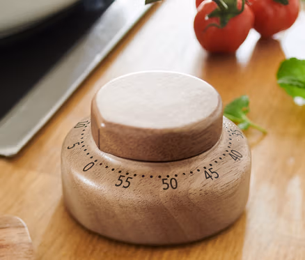 Auf einem Holztisch stehen eine Schüssel mit Essen, ein hölzerner Kurzzeitmesser, Cherrytomaten und ein Brett mit Feta und Blüten.