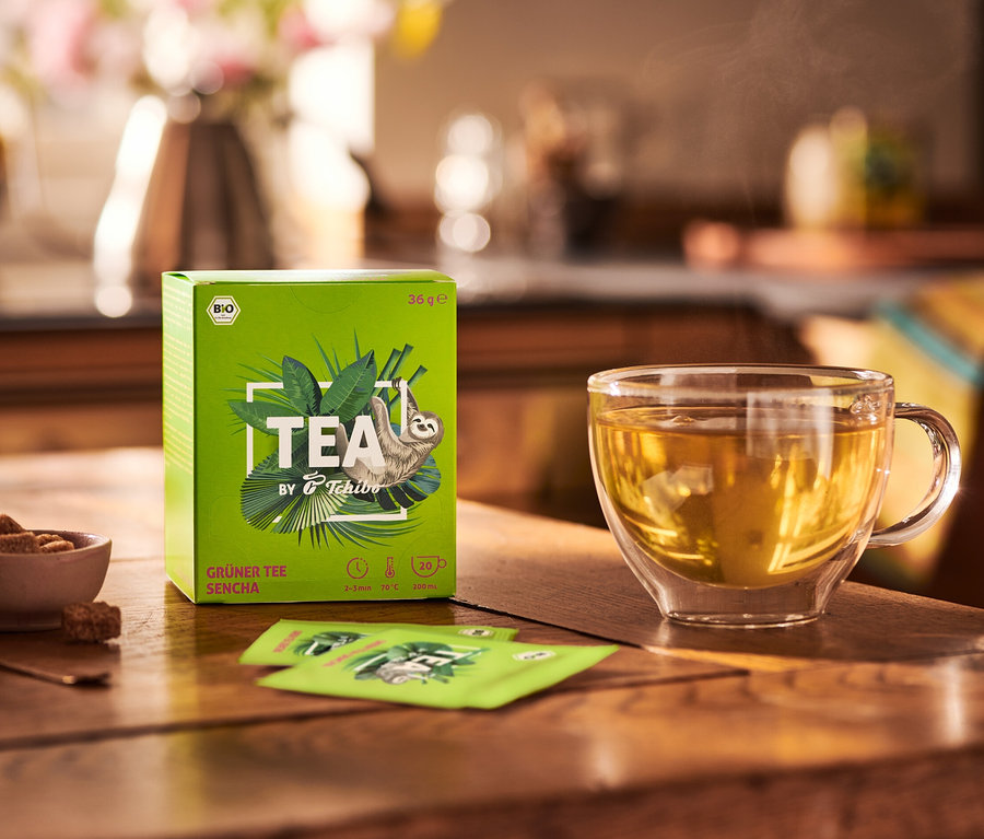 Auf einem Holztisch stehen eine Packung TEA by Tchibo Bio Grüner Tee Sencha - 8x 20 Teebeutel, eine Schale mit Zucker und eine Tasse Tee.