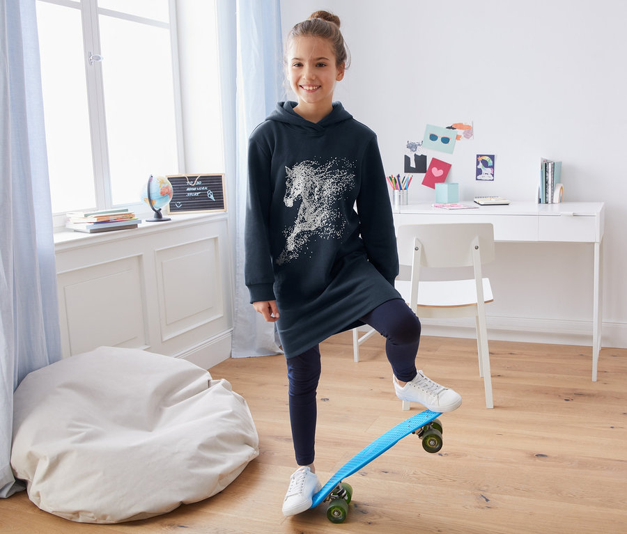 Mädchen steht mit einem Bein auf einem Skateboard und trägt ein Sweatkleid.