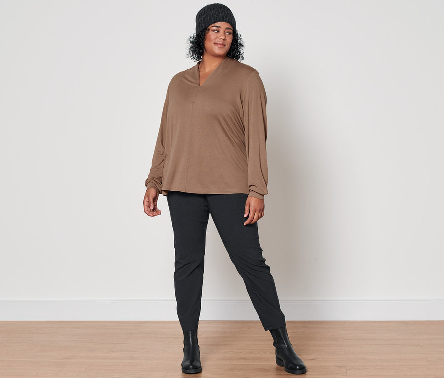 Eine Frau posiert in einem beigen Langarmshirt mit V-Ausschnitt, einer schwarzen Bengalinhose, Lederboots und einer anthrazitfarbenen Strickmütze mit Wolle.