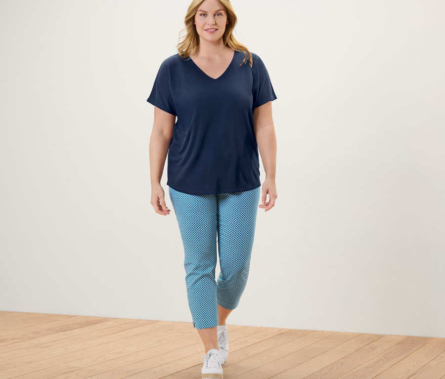 Frau geht und trägt eine 7/8-Stretchhose mit Alloverprint, ein T-Shirt mit V-Ausschnitt und Turnschuhe.