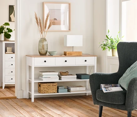 Wohnzimmer Interieur mit einem anthrazitfarbenen, gepolsterten Sessel, einer Anrichte mit Schubladen und einer Tischleuchte mit Holzfuß.