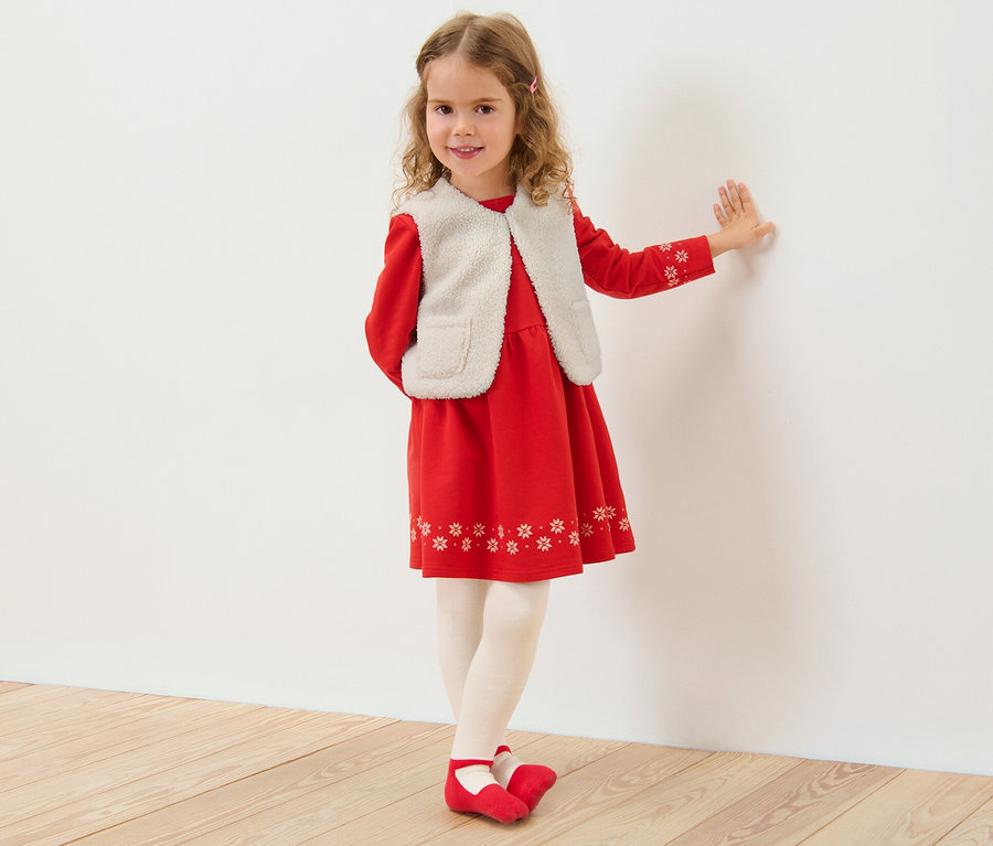 Mädchen lehnt an einer weißen Wand und trägt ein rotes Kleid und eine Kinder-Teddyweste.