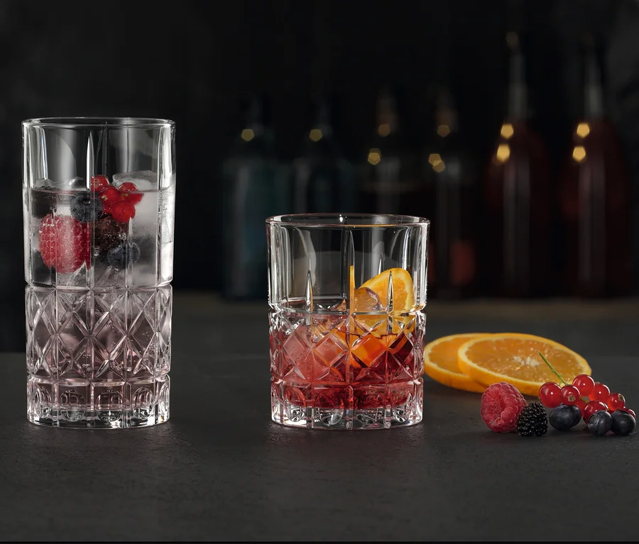 Zwei Nachtmann Gläser, ein Longdrinkglas »Highland« mit Beeren und Eis, ein Tumbler »Highland« mit Orangen und Eis, auf dunkler Fläche.