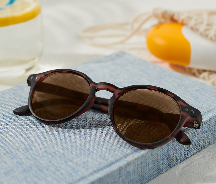 Getönte Lesebrille in Braun liegt auf einem blauen Buch. Im Hintergrund ein Glas Wasser mit Zitrone und eine Sonnencreme in einer Tasche.