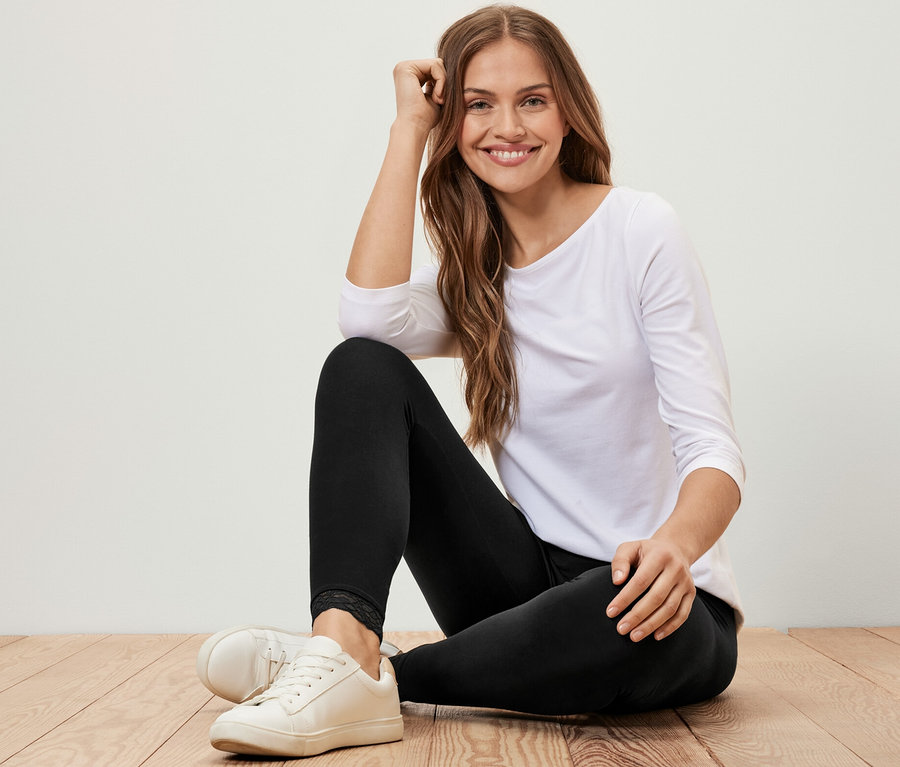 Eine Frau sitzt auf einem Holzboden und trägt Leggings mit Spitzenborte, ein weißes Dreiviertelarmshirt und weiße Turnschuhe.